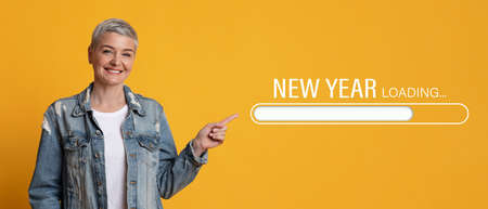 Smiling mature woman pointing finger aside at New Year loading process bar on orange studio background, collageの写真素材
