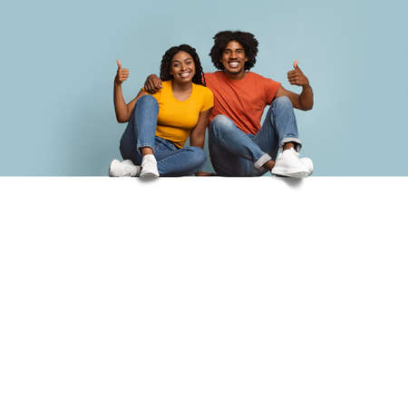 Positive black couple sitting on empty horizontal board for advertisement