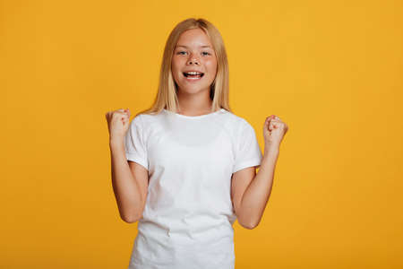Happy excited teenager blonde girl pupil in white t-shirt rejoices victory and gestures