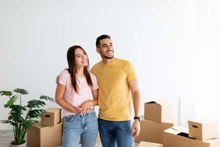 Happy young multiracial couple standing in their house among cardboard boxes on moving day, copy...