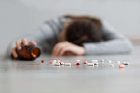 Unhappy european millennial woman lying on floor, focus on scattered pills at home, blurredの写真素材