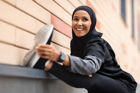Smiling Sporty Muslim Woman In Hijab Stretching Leg Muscles Before Training Outdoorsの写真素材