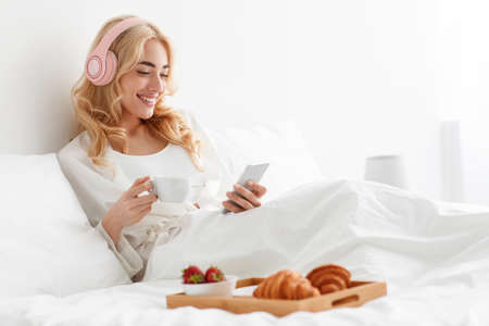 Smiling millennial pretty european blonde woman in robe and headphones reading news, chatting, watch videoの写真素材