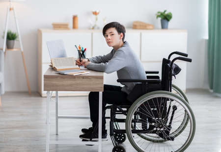 Disabled teenager in wheelchair learning online with books and laptop computer at homeの写真素材