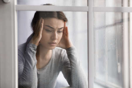 Upset female suffering from health problems, depression and headache, near window with dropsの写真素材