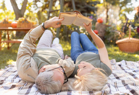 Married senior couple reading book together while lying on picnic blanket in garden, spending free time outdoorsの写真素材
