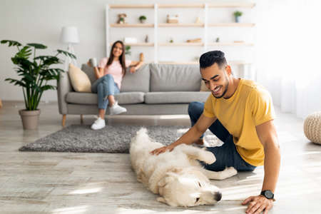 Happy Arab guy stroking cute dog on floor at home, his girlfriend relaxing with cup of coffee on background, copy spaceの写真素材