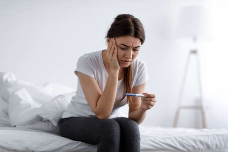 Depressed sad unhappy young pretty european woman looking at negative home test sitting on bed in bedroom interior aloneの写真素材