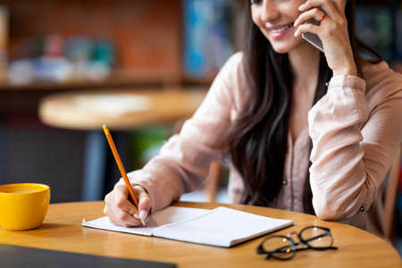 Looking for jop opportunity. Young woman talking on smartphone and taking notes, sitting at cafe interiorの写真素材