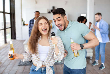 Young interracial couple singing song together into microphone, holding beer bottles, partying with friends at homeの写真素材