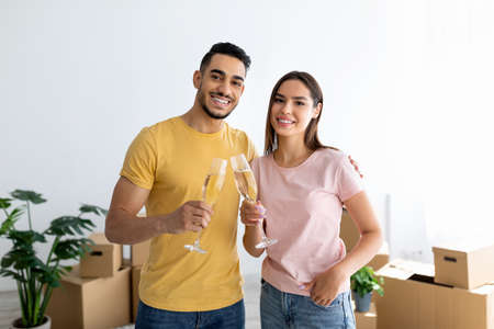 Loving interracial spouses celebrating moving day with champagne, toasting in new apartment among...