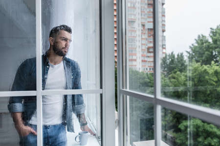 Depressed sad man looking through window, hoping for end of pandemicの写真素材