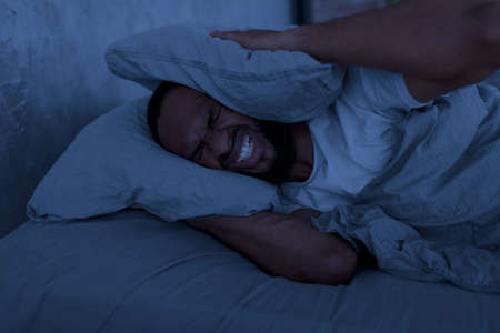 Stressed black man covering ears lying in bed at hightの写真素材