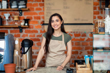 Opening small business. Happy latin lady in apron posing near bar counter and smiling to camera, waiting for clientsの写真素材
