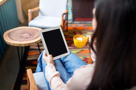 Modern technologies concept. Young woman using digital tablet with blank screen and drinking coffee, sitting in cafeの写真素材