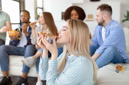 Pretty young blonde lady eating pizza on home party with her multiethnic friends, free spaceの写真素材