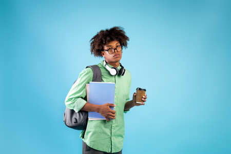 Exhausted male student with notebooks, takeaway coffee and backpack looking at camera on blue studio backgroundの写真素材