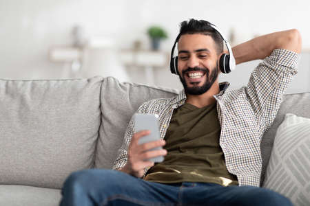Smiling young Arab guy in headphones enjoying music or audio book on smartphone, picking popular playlist at homeの写真素材