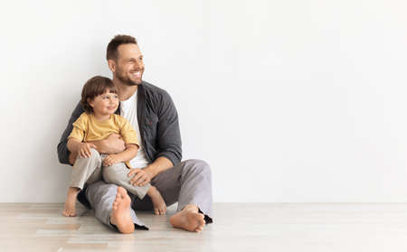 Happy caucasian father carrying little son, sitting together on floor and looking aside at empty space, white backgroundの写真素材