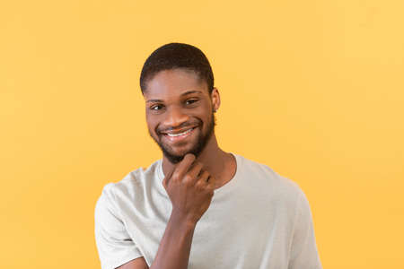 Cheerful african american guy touching chin, smiling and looking at camera while posing over yellow backgroundの写真素材