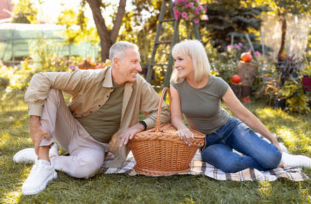 Lovely senior couple having picnic outdoors, sitting on blanket and smiling to each other, enjoying warm autumn eveningの写真素材