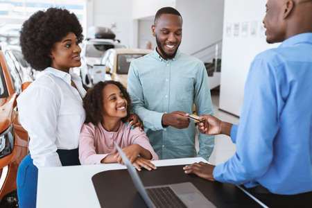 Cheerful black family paying for new car with credit card, making deal with salesman at auto showroomの写真素材