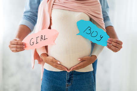 Young muslim couple holding BOY and GIRL cards near pregnant belly, guessing their future babys genderの写真素材
