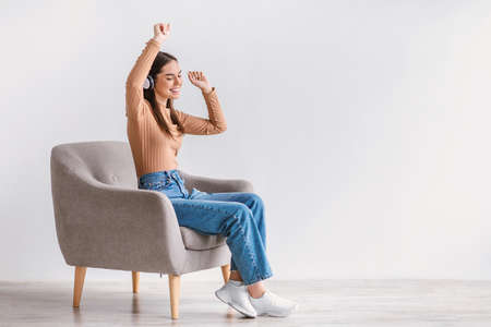 Pretty young lady in headset sitting in armchair, listening to music, lifting hands up, dancing to...
