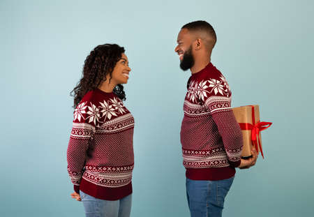 Surprise for Christmas. Black guy holding gift box behind back, congratulating wife over blue studio backgroundの写真素材