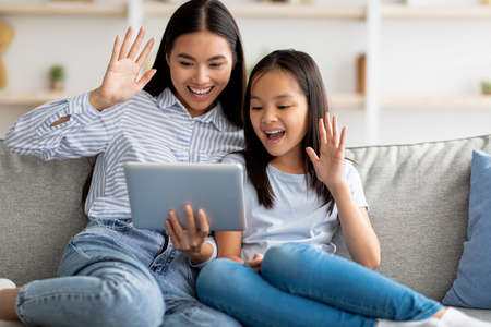 Happy asian mother and daughter having video call on digital tablet, sitting on sofa and waving at...