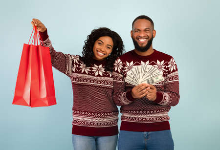 Excited african american spouses in Christmas sweaters holding shopping bags and bunch of money, blue backgroundの写真素材