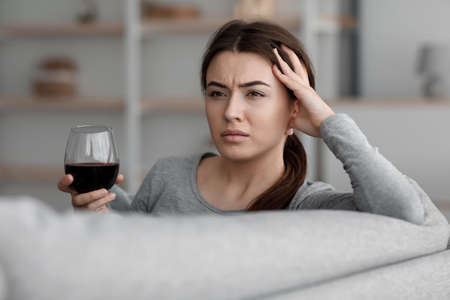 Drunk unhappy caucasian young woman with glass of wine suffering from depression and headache and relax at homeの写真素材