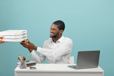 Young black office worker receiving pizza delivery from courier, sitting at workplace over blue backgroundの写真素材