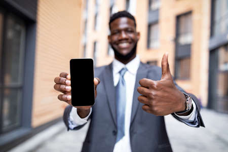 Happy black businessman showing smartphone with blank screen and gesturing thumb up, selective focusの写真素材