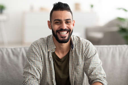 Portrait of attractive young Arab man smiling and looking at camera, sitting on couch at homeの写真素材