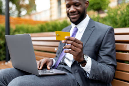 Young african american businessman using credit card and laptop, doing some online shopping, sitting outdoorsの写真素材