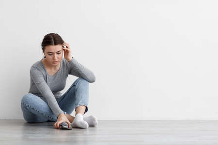 Upset worried european millennial lady suffering from depression sitting on floor and taking smartphone at homeの写真素材