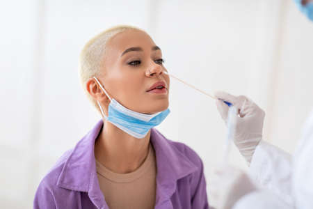 Coronavirus diagnostic. Nurse making covid-19 PCR test for young lady using sterile swab stick, sitting over white wallの写真素材