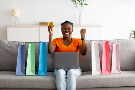 Young black woman with credit card and laptop buying via internet at home, making YES gesture. Online shoppingの写真素材