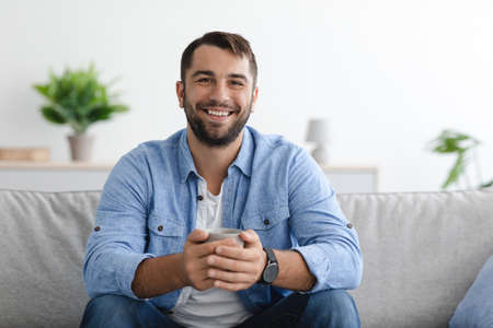 Cheerful middle aged caucasian man with beard in casual hold cup of drink look at camera at homeの写真素材