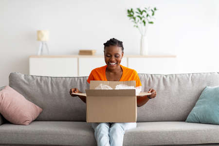 Cheery young black woman opening carton box, satisfied with delivery service, receiving online store order at homeの写真素材