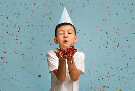 Happy Birthday. Little Asian Boy Wearing B-Day Hat Blowing Confetti At Camera