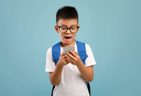 Excited Asian Schoolboy With Backpack Using Smartphone Over Blue Backgroundの写真素材