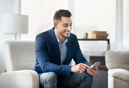 Smiling Arab businessman in formal wear using mobile phone, checking business messages, sitting in armchair at officeの写真素材