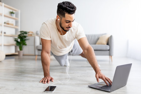 Young Arab guy doing plank, wearing headphones, listening to music, following online video tutorial on laptop at homeの写真素材