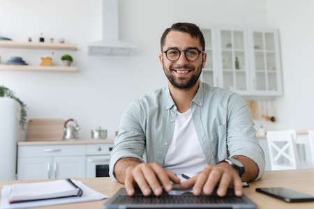 Cheerful mature caucasian male with beard in glasses typing on keyboard, work on laptop, look at webcamの写真素材