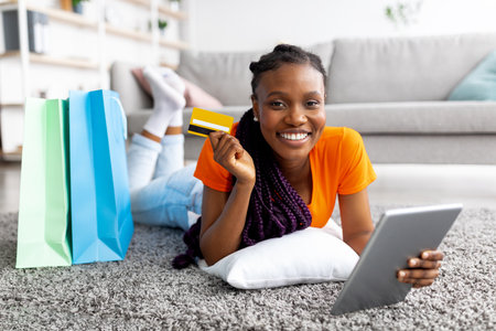 Beautiful smiling black lady lying on floor with digital tablet, credit card and shopping bags at homeの写真素材
