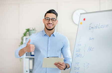 Positive Arab male teacher showing thumb up near blackboard, recommending new online foreign languages schoolの写真素材