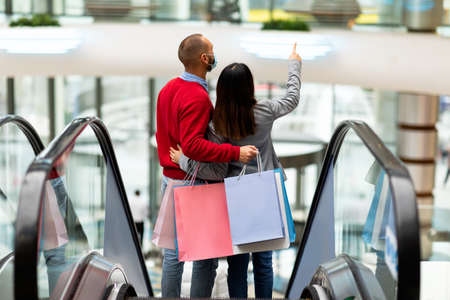 Young international couple in face masks standing on moving stairway, shopping together in city mall during covidの写真素材