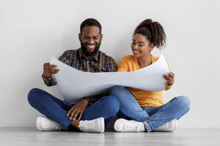 Smiling young black couple looking at apartment plan on big paper sitting on floor in empty roomの写真素材